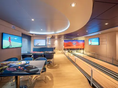 Die Sportsbar und Bowlingbahn an Bord der MSC Preziosa. Links Tische, einer mit Rennwagenmotiv. Mehrere Bildschirme zeigen Tennis und ein Highway 66. Rechts zwei Bowlingbahnen.