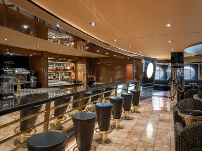 Die elegante Bar an Bord der MSC Preziosa zeigt eine lange Bartheke mit Hockern, eine Kaffeemaschine, Spirituosenregale und im Hintergrund gemütliche Lounge-Bereiche mit Sofas und Sesseln.