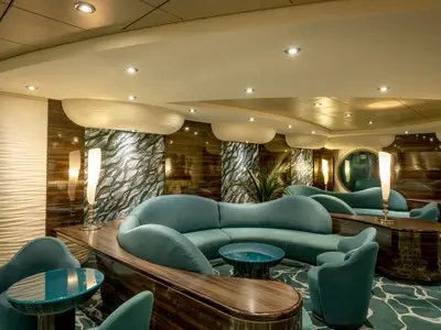 Elegante Bar-Lounge an Bord der MSC Preziosa. Geschwungene türkisfarbene Sofas, dunkle Holzmöbel und dekorative Wände. Warme Beleuchtung schafft gemütliche Abendstimmung.