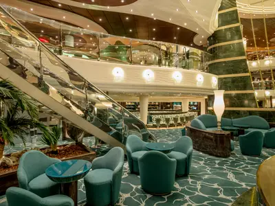 Elegante Bar-Lounge an Bord der MSC Preziosa. Im Atrium befinden sich eine geschwungene Treppe, bequeme Sessel und eine beleuchtete Bar. Grüne Marmorwände und Palmen.