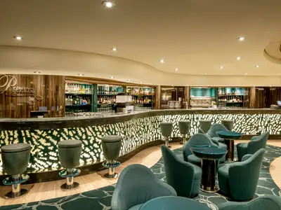 Die Preziosa Bar an Bord der MSC Preziosa. Elegante Theke mit beleuchtetem grünem Muster, Barhockern und Lounge-Sesseln. Gut bestückte Spirituosenregale im Hintergrund.