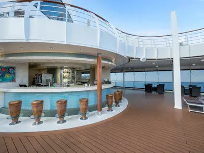 An outdoor bar area named Turquoise on the MSC Preziosa cruise ship deck, featuring a curved bar with wooden stools and a panoramic sea view. Lounge seating is visible further back.