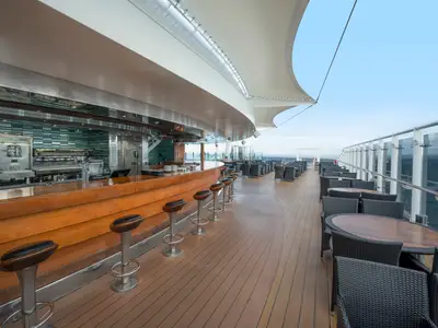 Panoramablick auf die teilweise überdachte "The Garden Bar" auf dem Oberdeck der MSC Preziosa. Ein langer Holztresen mit Barhockern und zahlreiche Tische mit Flechtstühlen laden zum Verweilen ein. Im Hintergrund das weite Meer.
