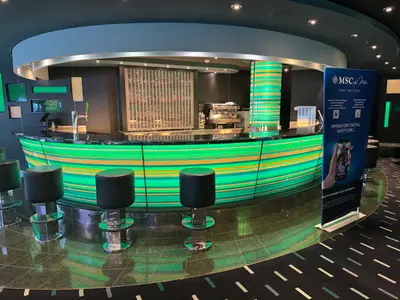 Eine modern gestaltete Bar an Bord der MSC Preziosa mit auffällig grün-gelb gestreifter Front, Barhockern und Info-Schild.