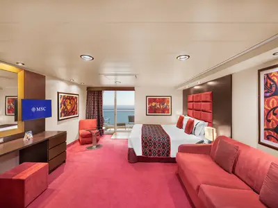 Blick in eine barrierefreie Balkonkabine an Bord der MSC Preziosa. Ein Doppelbett, ein rotes Sofa und eine Kommode sind zu sehen. Vom Balkon aus ist das Meer sichtbar. Rote Farbe dominiert die Einrichtung und Kunstwerke.