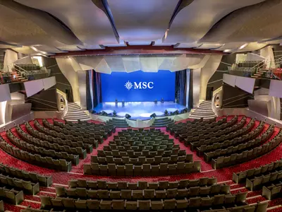 Das leere Platinum Theatre an Bord der MSC Preziosa mit rotem Teppich, dunklen Sitzen, einer Bühne mit blauem MSC-Logo und geschwungenen Treppen.