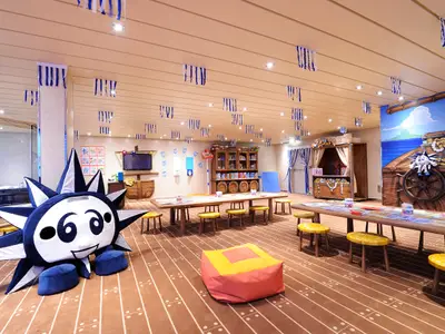 Großer, heller Kinderbereich Doremiland an Bord der MSC Preziosa mit vielen kleinen Tischen, Stühlen und einem großen Sonnen-Maskottchen als Sitzgelegenheit. Piraten-Themenwandbild am Ende des Raumes.