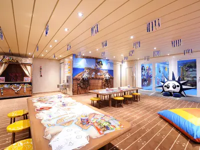 Ein bunter Kinderbereich an Bord der MSC Preziosa mit einem Piraten-Wandgemälde, Basteltischen, gelben Hockern und einem Puppentheater. Ein großer schwarzer Kuschel-Stern steht vor den Glastüren.