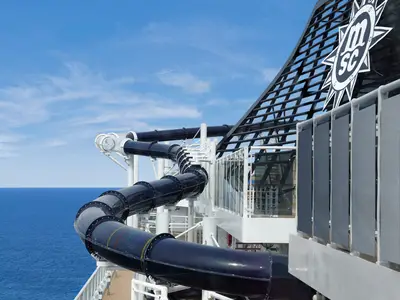 Die geschwungene, dunkelblaue Wasserrutsche Vertigo auf der MSC Preziosa schlängelt sich über das Sonnendeck mit blauem Himmel und offenem Meer im Hintergrund. Rechts ist die obere Struktur des Schiffes mit einem MSC Logo sichtbar.