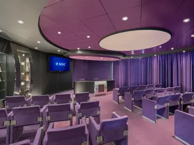 Moderner Konferenzraum an Bord der MSC Preziosa mit violetten Samtsesseln. Ein Podium, Bildschirm mit MSC Logo, lila Vorhänge und stilvolle Deckenbeleuchtung definieren den Raum.