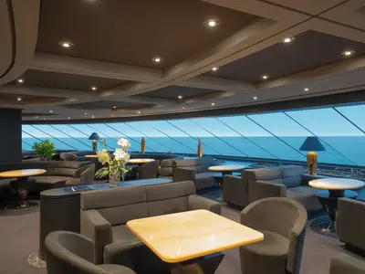 Weitwinkelaufnahme der eleganten Top Sail Lounge an Bord der MSC Preziosa. Komfortable Sitzbereiche mit dunklen Sofas und Sesseln, hellen Tischen. Große Fensterfront bietet Panoramablick auf das Meer.