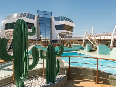 MSC Preziosa: Der Aqua Park mit mehreren Pools und dekorativen Kakteen-Skulpturen. Im Hintergrund die moderne Glas-Architektur des Schiffes. Wenige Gäste entspannen auf dem sonnigen Pooldeck.