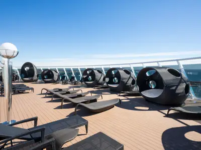 Das sonnige Sonnendeck der MSC Preziosa zeigt schwarze Rattan-Lounge-Sessel und kugelförmige Sitzinseln auf einem Holzdeck. Eine weiße Laterne ragt in den Vordergrund, im Hintergrund ist das Meer und ein blauer Himmel zu sehen.