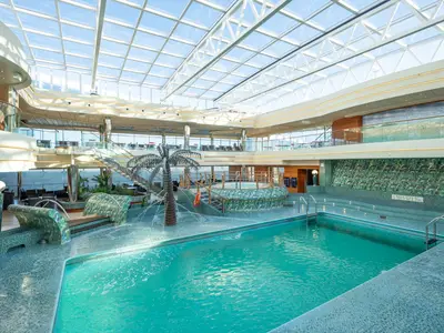 Heller La Playa Preciosa Poolbereich an Bord der MSC Preziosa. Mit großem Pool, Whirlpool, künstlichem Wasserfall und Glasdach. Obere Decks mit Lounge-Sitzplätzen sichtbar.