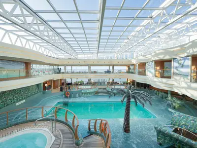 Weitwinkelaufnahme des hellen, überdachten Poolbereichs La Playa Preciosa an Bord der MSC Preziosa mit einem großen Pool, mehreren Whirlpools, einer Palmenstatue und Mosaik-Verzierungen unter einem Glasdach. Das Deck ist ohne Personen.