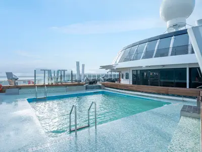Blick auf das Hauptdeck der MSC Preziosa mit einem grossen Aussenpool und Jacuzzi, umgeben von Mosaikfliesen und Holzdeck. Im Hintergrund gläserne Aufbauten und das offene Meer.