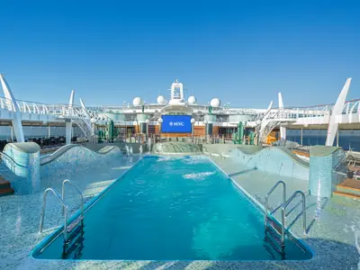 Ein Weitwinkelbild des Acqua Parks an Bord der MSC Preziosa zeigt einen großen, türkisblauen Pool umgeben von einem mosaikverzierten Wellenbereich. Am Ende des Pools thront eine riesige LED-Leinwand mit dem MSC-Logo. Im Hintergrund sind Teile der Schiffsarchitektur, Sonnenliegen und grüne Kakteen-Skulpturen unter einem klaren blauen Himmel zu sehen. Der Pool ist leer und das Wasser sprudelt an einigen Stellen.