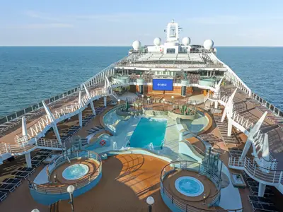 Vogelperspektive auf den Aqua Park der MSC Preziosa mit mehreren Pools, Whirlpools, Liegestühlen und einem großen LED-Bildschirm. Das Schiff fährt auf blauem Meer unter klarem Himmel.