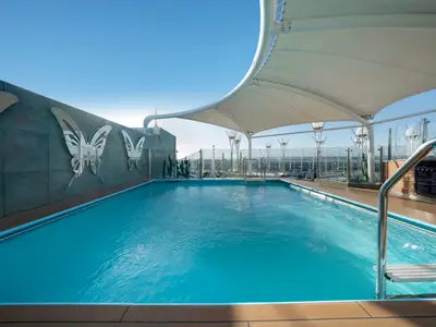 Der Poolbereich "The One Pool" an Bord der MSC Preziosa. Türkisblaues Wasser, Holzdeck, spiegelnde Schmetterlinge an der Wand, Sonnensegel und blauer Himmel.