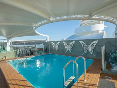 Aussenpool auf der MSC Preziosa. Blauer Pool, Holzdeck, weisses Sonnendach. Drei grosse, silberne Schmetterlinge dekorieren die Wand.