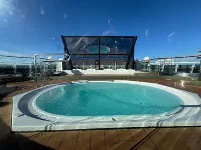 Ein runder Whirlpool mit türkisblauem Wasser auf dem hölzernen Sonnendeck an Bord der MSC Preziosa unter klarem blauem Himmel.