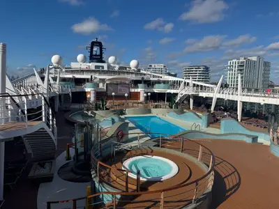Blick über das sonnige Pooldeck an Bord der MSC Preziosa mit einem runden Whirlpool, dem Hauptpool und einer Stadt im Hintergrund.
