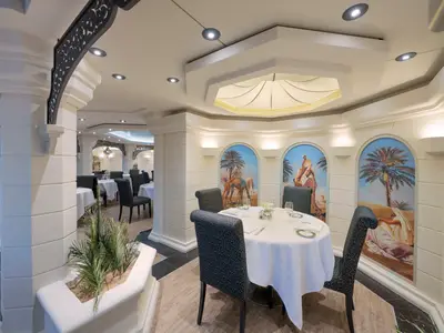 Blick in das elegant gestaltete La Palmeraie Restaurant an Bord der MSC Preziosa. Mehrere runde Tische sind festlich mit weißen Tischdecken und Geschirr gedeckt. Die Wände zieren bogenförmige Wandmalereien, die orientalische Wüstenszenen mit Palmen, Kamelen und Figuren zeigen. Säulen und indirekte Beleuchtung prägen das Interieur.