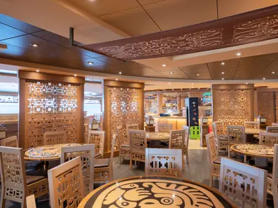 Ansicht des "Inca & Maya" Buffet-Restaurants an Bord der MSC Preziosa. Der Raum ist mit hellem Holz, kunstvollen Trennwänden und runden Tischen mit gemusterten Glasplatten ausgestattet. Im Hintergrund sind Fenster mit Meerblick und eine Buffettheke zu sehen. Keine Personen anwesend.