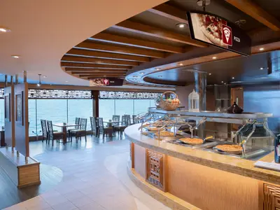 Ein helles Buffet-Restaurant an Bord der MSC Preziosa mit großen Fenstern zum Meer. Mehrere Pizzen liegen auf einer Theke bereit, dahinter Tische und Stühle für Gäste.