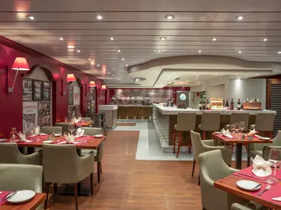 Innenansicht des Spezialitätenrestaurants Butcher's Cut an Bord der MSC Preziosa. Mehrere Tische mit Stühlen, rote Wände mit Lampen und eine offene Küche mit Bar sind sichtbar.
