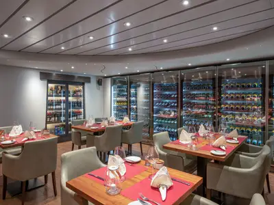 Das elegante Restaurant an Bord der MSC Preziosa zeigt mehrere Tische mit rotem Tischset und beige Stühlen. Entlang der Wand erstreckt sich eine beleuchtete Weinwand mit zahlreichen Flaschen.