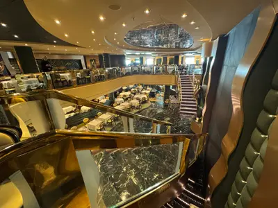 Blick von einer goldenen Treppe auf das zweistöckige Hauptrestaurant an Bord der MSC Preziosa mit gedeckten Tischen und glänzendem Boden.