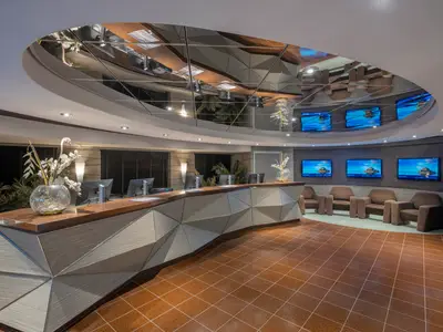 Die helle, moderne Rezeption an Bord der MSC Preziosa zeigt eine lange, geschwungene Theke mit Arbeitsplätzen und Bildschirmen. Ein gefliester Boden und eine Sitzgruppe mit bequemen Sesseln sind ebenfalls zu sehen. Die Decke ist teilweise verspiegelt.