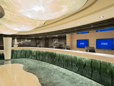 Moderne Rezeption der MSC Preziosa mit geschwungener Theke aus grünem Marmor. Monitore zeigen das MSC-Logo und 'Preziosa guest service' ist zu lesen. Wellenförmige Decke und gemusterter Boden.