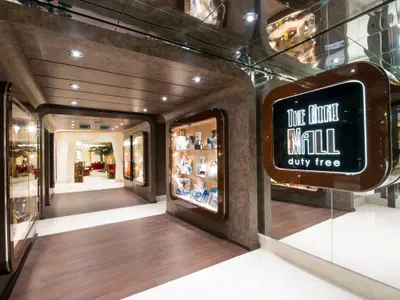 Der elegante Duty-Free-Einkaufskorridor „The Mini Mall“ an Bord der MSC Preziosa mit beleuchteten Vitrinen, einem Leuchtschild und einer Lounge im Hintergrund.