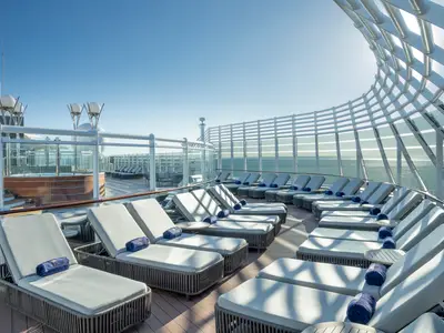 Ein sonniges Solarium-Sonnendeck der MSC Preziosa mit vielen Liegestühlen, einem Whirlpool und Meerblick durch eine moderne Glas-Metall-Struktur. Blaue Handtücher liegen auf den Liegen. Der Himmel ist klarblau.