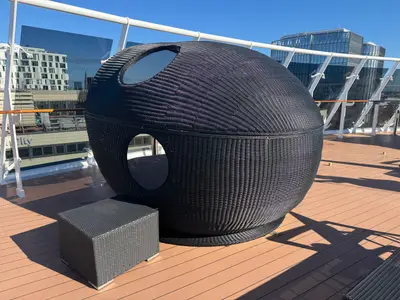 Ein runder, schwarzer Korb-Liegestuhl auf dem hölzernen Sonnendeck an Bord der MSC Preziosa mit Blick auf eine Skyline.