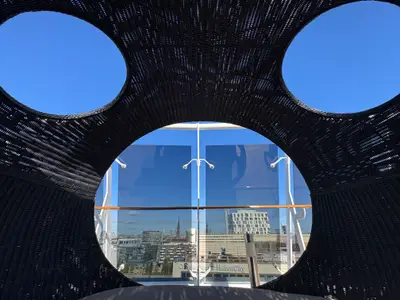 Blick durch ein rundes Korbgeflecht auf dem Sonnendeck der MSC Preziosa auf eine verglaste Brüstung und eine Stadt im Hintergrund.