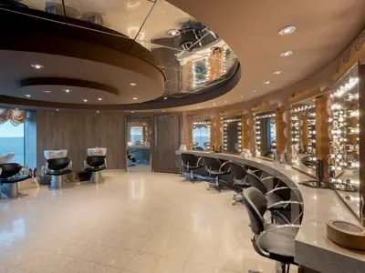 Ein moderner Friseursalon an Bord der MSC Preziosa. Links stehen Friseurstühle mit Waschbecken vor einem großen Fenster mit Meerblick. Rechts eine geschwungene Theke mit vielen beleuchteten Spiegeln.