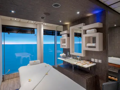 Ein heller, moderner Massageraum an Bord der MSC Preziosa mit raumhohen Fenstern und weitem Blick auf das blaue Meer und den Horizont. Eine Massageliege, Handtücher mit "MSC Aurea Spa"-Logo, ein Waschbecken und Holzutensilien sind sichtbar.