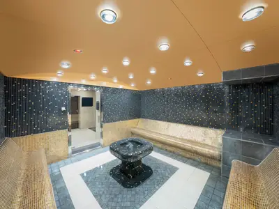 Ein luxuriöser Thermalbereich im MSC Aurea Spa an Bord der MSC Preziosa mit dunklen und goldenen Mosaikfliesen, geschwungenen Sitzbänken und einem zentralen Brunnen. Die Decke ist goldfarben mit vielen Einbaulampen.
