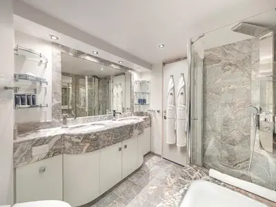 Helles Badezimmer einer Suite an Bord der MSC Preziosa mit Doppelwaschbecken aus Marmor und einem großen Spiegel. Eine moderne Dusche mit Regenduschkopf und Handbrause ist zu sehen. Zwei weiße Bademäntel hängen an der Tür, und auf Glasregalen stehen Pflegeprodukte bereit.
