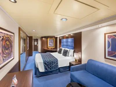 Eine geräumige Premium Suite Aurea an Bord der MSC Preziosa. Sie zeigt ein großes Doppelbett mit blauer Tagesdecke, ein passendes Sofa, einen Schreibtisch und Kunstwerke an den Wänden. Die Suite ist in Blautönen und Holzakzenten gehalten und strahlt Gemütlichkeit aus.