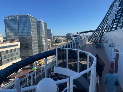 Außendeck an Bord der MSC Preziosa mit der großen blauen Wasserrutsche Vertigo und Blick auf moderne Glasgebäude im Hafen.
