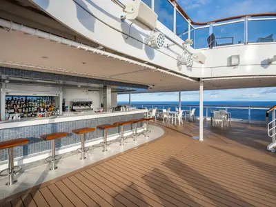Aussenansicht der Marina Bar an Bord der MSC Seascape: Eine moderne Bar mit Theke, Hockern und vielen Flaschen. Dahinter ein Deck mit Tischen und dem weiten blauen Ozean.