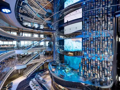 Die mehrstöckige Atrium-Lobby der MSC Seascape zeigt eine futuristische Architektur mit gläsernen Treppen, Spiegeln und einer zentralen Bühne. Ein großer Bildschirm zeigt Meerwellen. Verschiedene Bars und Lounges sind auf den Ebenen verteilt.