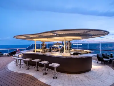 Die elegant beleuchtete Outdoor-Bar "Sky Bar" auf dem Deck der MSC Seascape zur Dämmerung. Mit Hockern, Loungemöbeln und Meerblick unter blauem Himmel.
