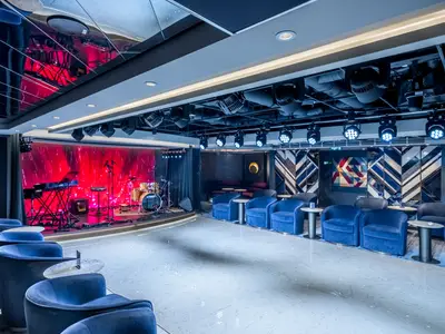 Eine Bühne mit Schlagzeug und Keyboard sowie blaue Samtsessel mit Tischen in der Uptown Lounge an Bord der MSC Seascape. Eine Wand ist mit rotem Licht und Mustern beleuchtet, die andere mit geometrischen Holzintarsien.
