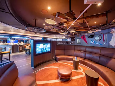 Eine breite Ansicht der Sports Bar an Bord der MSC Seascape mit einer runden Theke, Barhockern und einer halbkreisförmigen Ledersitzgruppe vor einem Fernseher. Die Wände sind mit Sportmemorabilien wie Baseballschlägern, Tennisschlägern, einem Trikot und Wimpeln dekoriert. Der Boden ist mit einem orangefarbenen Teppich in Form eines Basketballfeldes versehen. Die Szene ist beleuchtet, ohne Personen.