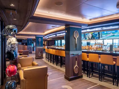 Blick in die Sports Bar an Bord der MSC Seascape mit einer langen Bar, goldenen Barhockern und grossen Bildschirmen. An den Säulen hängen Tennisschläger, und im Vordergrund sind Sporthelme ausgestellt. Keine Personen sichtbar.